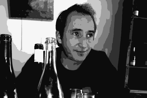 Luc Devot in black and white, looking toward the camera at a table with wine bottles