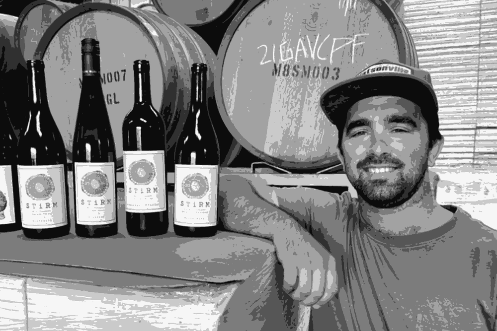 Ryan Stirm of Stirm Wine Co. smiling alongside a lineup of Stirm bottles in his Watsonville winery