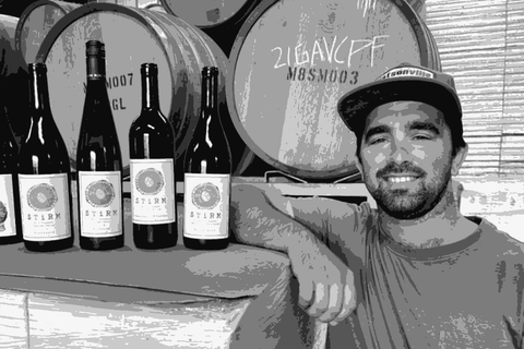 Ryan Stirm of Stirm Wine Co. smiling alongside a lineup of Stirm bottles in his Watsonville winery