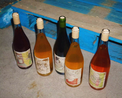 Five multi color bottles of natural wine on a concrete floor in front of a blue pallet at Primal Wine natural wine store - primalwine.com